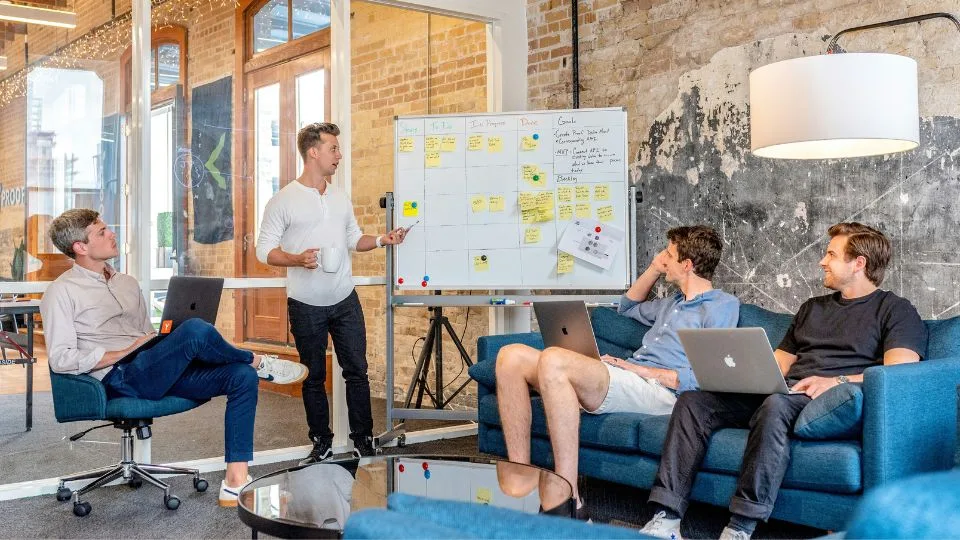 4 männliche Team Mitglieder, die im einem Raum teilweise auf einer Couch sitzen und an einer Pinwand im Rahmen eines Team Boost Workshops diskutieren.
